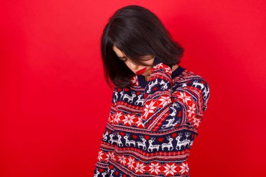 Esmer, beyaz kadın kırmızı arka planda Noel süveteri giyiyor boynu ağrıyor, boynuna elle dokunuyor, kas ağrısı çekiyor.