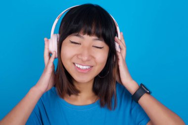 Mavi arka planda mavi t-shirt giyen genç Asyalı kadın gülümsüyor. En sevdiği müziği kablosuz kulaklıkla dinlediği için çok mutlu. Gözlerini kapatıyor..