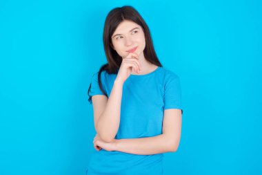 Mavi tişört giyen düşünceli genç beyaz kız mavi stüdyo arka planında izole edilmiş çenesini kapalı tutuyor ve büyük bir plan yapıyor.