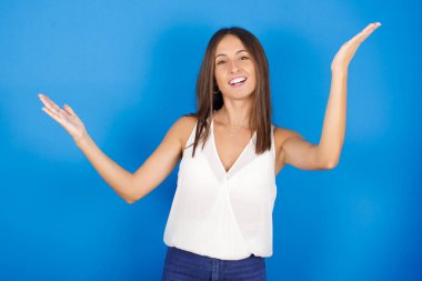 Neşeli, güzel genç bir kadın hoş geldin jesti yapıyor kollarını başının üstüne kaldırıyor..