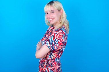 Mavi arka planda izole edilmiş çiçek desenli elbise giyen beyaz bir kızın portresi kollarını kavuşturmuş ve gülümsüyor.