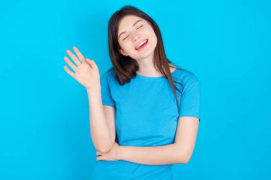 Mavi tişört giyen başarılı beyaz kız mavi stüdyo arka planına izole edilmiş olarak avucunu kaldırıyor ve arkadaşları tarafından eğlendirilirken gözlerini kapıyor.