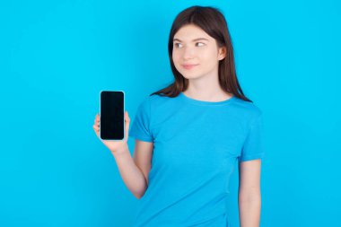 Mavi tişört giyen genç beyaz kız mavi stüdyo arka planında izole edilmiş yeni bir cep telefonu tutuyor ve gizemli görünüyor.