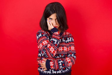 Çok sıkılmış esmer, beyaz bir kadın kırmızı arka planda Noel süveteri giyiyor, el ele tutuşuyor ve diğer eli çapraz tutuyor, yorgun ve hasta görünüyor.,