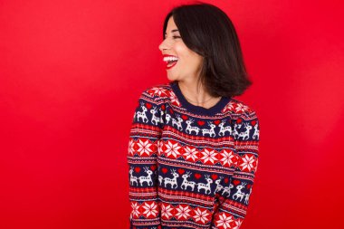 Esmer beyaz kadın kırmızı arka planda Noel süveteri giyiyor. Yeni planlar için çok mutlu ve heyecanlı..