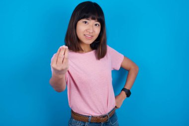 What the hell are you talking about. Shot of frustrated young asian woman wearing pink t-shirt against blue background gesturing with raised hand doing Italian gesture, frowning, being displeased and confused with dumb question.