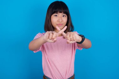 Mavi arka planda pembe t-shirt giyen genç Asyalı kadın olumsuz işaret veren kızgın bir ifadeye sahip..