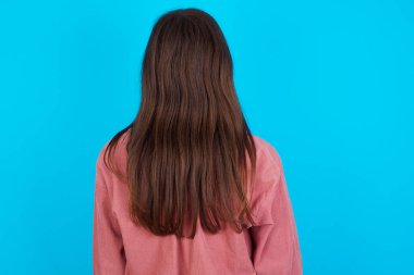 Pembe tişörtlü genç beyaz kız, mavi arka planda izole edilmiş. Arkaya bakıyor, kolları vücudunda..
