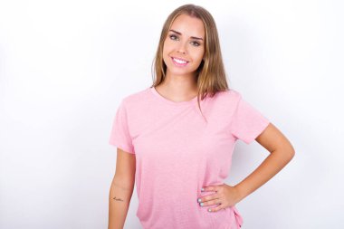 Beyaz arka planda pembe tişört giyen neşeli beyaz bir kızın stüdyo fotoğrafı elini kalçasında tutuyor, geniş bir şekilde gülümsüyor.