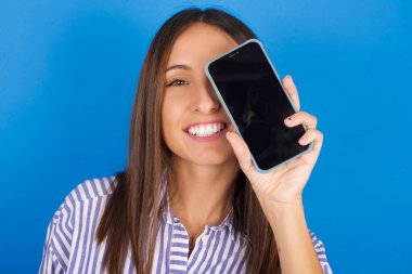 Modern akıllı telefonu tutan güzel genç bir kadın bir gözünü kapatıp gülümsüyor.