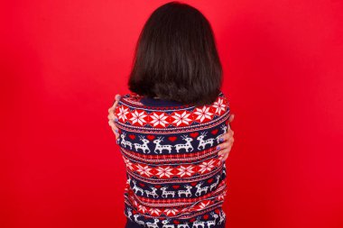 Esmer, beyaz bir kadın, kırmızı arka planda Noel süveteri giyiyor, mutlu ve pozitif bir şekilde kendini kucaklıyor. Sevgi ve özgüven.