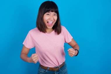 Neşeli, şanslı Asyalı kadın pembe tişört giyiyor mavi arka planda tezahürat yapıyor, başarıyı kutluyor, yumruklarını sıkarak evet diye bağırıyor.