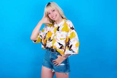 Mavi arka plan üzerine izole edilmiş çiçekli gömlek giyen beyaz kız telefon konuşmasını taklit ediyor, telefonla el hareketi yapıyor, kendine güvenen bir ifadesi var. Ara beni.!