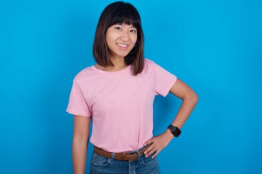 Mavi arka planda pembe tişört giyen neşeli Asyalı kadının stüdyo fotoğrafı elini kalçasında tutuyor, geniş bir şekilde gülümsüyor.