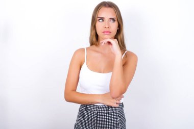 Beyaz arka planda beyaz kolsuz bluz giyen düşünceli genç beyaz bir kız elini çenenin altında tutuyor, düşünceli bir şekilde yukarı bakıyor..