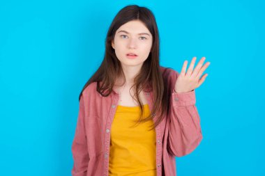 Studio shot of frustrated young caucasian girl wearing pink shirt isolated over blue background gesturing with raised palm, frowning, being displeased and confused with dumb question.