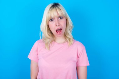 Etkileyici yüz ifadeleri. Şok olmuş beyaz kız pembe tişört giyiyor. Mavi arka planda izole edilmiş. Gördüklerinden sersemlemiş hissediyor..