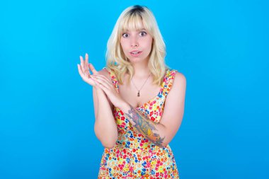 Mavi arka planda soyutlanmış çiçek desenli elbise giyen beyaz bir kız avuçlarını ovuşturur ve inançsızlıkla kameraya bakar.