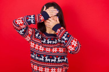 Esmer, beyaz kadın kırmızı arka planda Noel süveteri giyiyor gözleri ve ağzı kapalı, şaşırmış ve şok olmuş. Duyguları gizlemek.