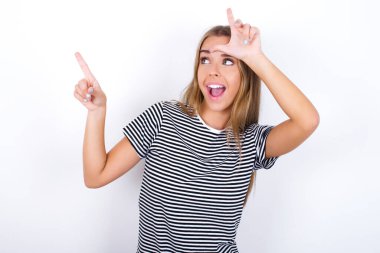 Beyaz arka planda çizgili tişört giyen güzel sarışın kız kaybeden işareti gösteriyor ve boş bir yeri işaret ediyor.