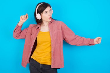Pembe gömlekli genç beyaz kız mavi arka planda izole edilmiş dans ediyor ve kulaklıklarla müzik dinliyor..