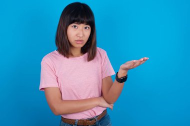 Studio shot of frustrated young asian woman wearing pink t-shirt against blue background gesturing with raised palm, frowning, being displeased and confused with dumb question.