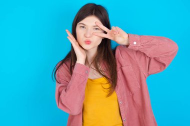 Mavi arka plan üzerinde izole edilmiş pembe tişört giyen genç beyaz kız gözlerinin yanına V işareti yapıyor. Boş hayat tarzı insanlar flört konseptini kutluyorlar..