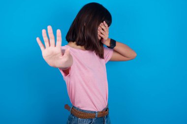 Mavi arka planda pembe t-shirt giyen genç Asyalı kadın avuçla gözlerini kapatıyor ve dur hareketi yapıyor, saklanmaya çalışıyor. Bana bakma, görmek istemiyorum, utanıyorum ya da korkuyorum..