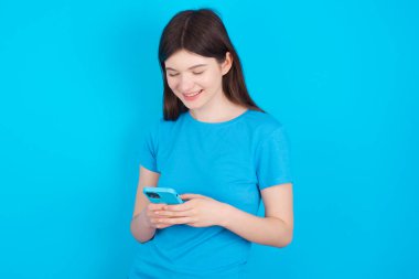 Mavi tişört giyen gülümseyen beyaz kız mavi stüdyo arka planında izole edilmiş cep telefonu kullanıyor, mesajlaşıyor, arkadaşlarıyla mesajlaşıyor, akıllı telefona bakıyor. Modern teknolojiler ve iletişim.