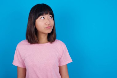 Mavi arka planda pembe t-shirt giyen genç Asyalı kadın düşünceli bir şekilde kenara bakıyor, üniversiteden sonra ne yapacağını düşünüyor..