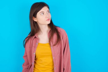 Mavi arka plan üzerinde izole edilmiş pembe tişört giyen genç beyaz kız düşünceli bir şekilde kenara bakıyor, üniversiteden sonra planlar yapıyor, yeni proje hakkında düşünüyor..