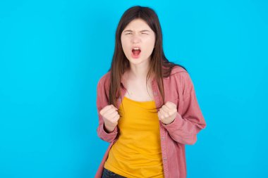 Neşeli, şanslı, pembe tişörtlü beyaz kız mavi arka planda izole edilmiş tezahürat yapıyor, başarıyı kutluyor, yumruklarını sıkarak evet diye bağırıyor.