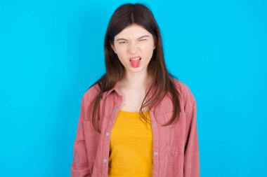 Vücut dili. Mide bulandırıcı, pembe gömlek giyen, mavi arka planda soyutlanmış, kaşlarını çatan, bir şeye karşı hoşnutsuzluk gösteren beyaz bir kız..