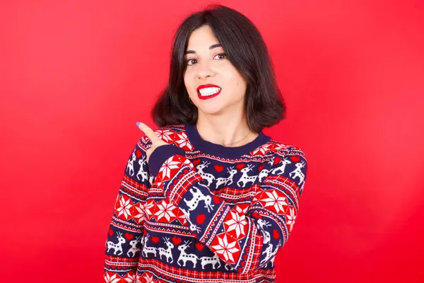 赤い背景にクリスマスセーターを着用しているブルネットの慎重な女性は 心配し 懸念とサプライズコンセプトと緊張を指摘 — ストック写真