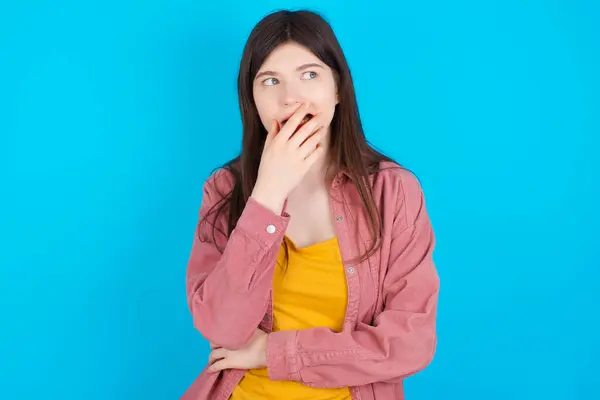 青い背景の上に隔離されたピンクのシャツを着ている若い白人少女は 何かまたは誰かが爪を噛むことを恐れた手で口を覆います — ストック写真