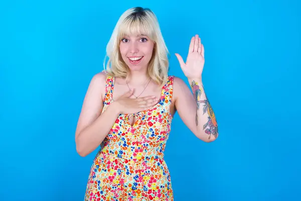 I swear, promise you not regret. Portrait of sincere caucasian girl wearing floral dress isolated over blue background raising one arm and hold hand on heart as give oath, telling truth, want you to believe.