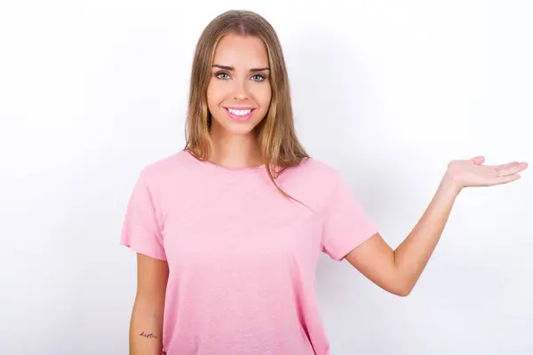 年轻的白人女孩穿着粉色T恤 背景是白色的 她很高兴地说 多么令人兴奋 有着令人惊奇的表情 用张开的手在空旷的空间里展示了一些东西 广告概念 — 图库照片