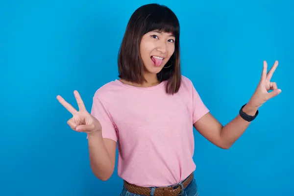 楽観的な笑顔で青い背景にピンクのTシャツを着た若いアジアの女性は 両手で平和や勝利のジェスチャーを示し 友好的に見えます Vサイン — ストック写真
