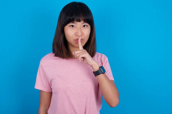 young asian woman wearing pink t-shirt against blue background makes hush gesture, asks be quiet. Don't tell my secret or not speak too loud, please!