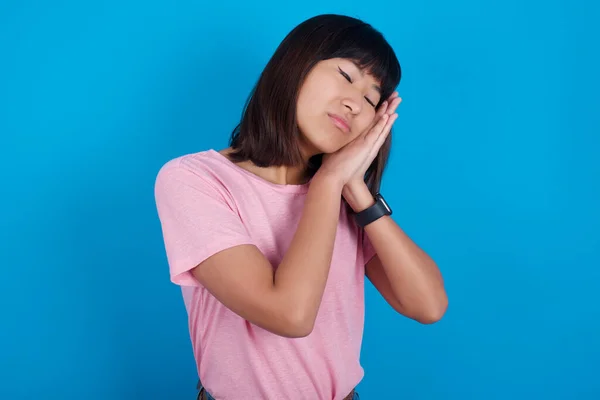 Relax and sleep time. Tired young asian woman wearing pink t-shirt against blue background with closed eyes leaning on palms making sleeping gesture.