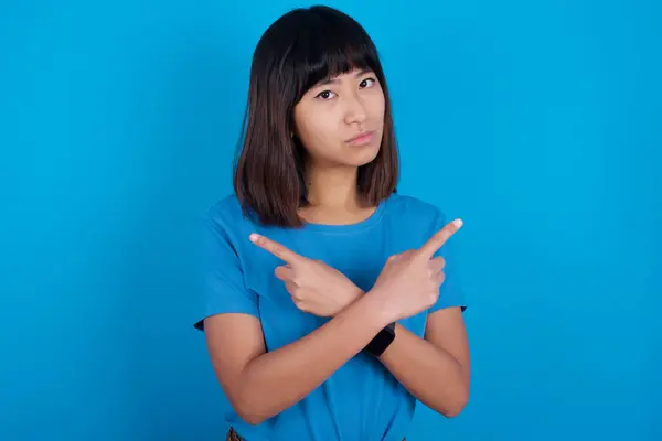 青い背景に青いTシャツを着ている真剣な若いアジアの女性は 2つのアイテムの間で躊躇する異なる側面で手とポイントを交差します ハード決定コンセプト — ストック写真