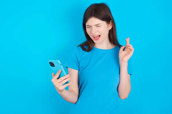 Photo of outraged annoyed young caucasian girl wearing blue t-shirt isolated over blue studio background  holds cell phone, makes call, argues with colleague,  expresses negative emotions. People and anger.