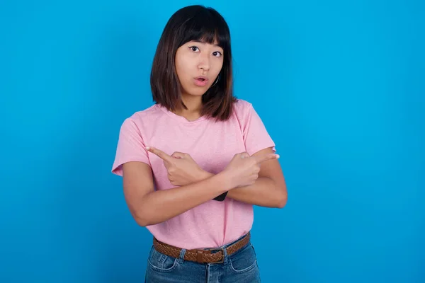 青い背景にピンクのTシャツを着ている混乱した若いアジアの女性は 2つの方法 交差した手で両側の点を選択し 疑問を感じます アドバイスが必要 — ストック写真