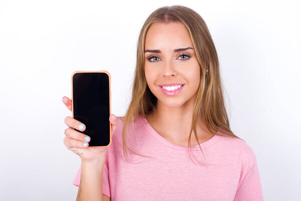 Улыбающаяся молодая белая девушка в розовой футболке на белом фоне показывает пустой экран телефона. Концепция рекламы и коммуникации.
