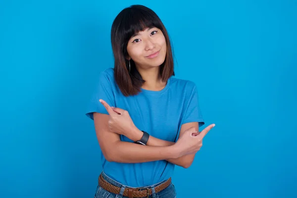 青い背景に青いTシャツを着ている若いアジアの女性は 両方のアイテムまたはバリアントの間で躊躇する異なる側面で腕とポイントを交差します 意思決定を支援する必要がある — ストック写真