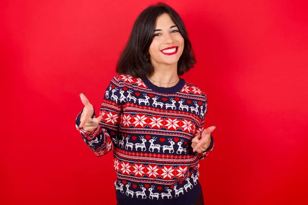 快乐的黑发女人 穿着圣诞毛衣 在红色背景下眨眼 用食指指着相机 选择某人 制造手枪 — 图库照片