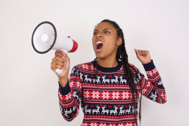 Beyaz duvara karşı Noel kazağı giyen Afro-Amerikalı kadın, elinde megafonla yüksek sesle bağırarak iletişim kuruyor, başarıyı ve olumlu konsepti ifade ediyor, pazarlama veya satış fikri.