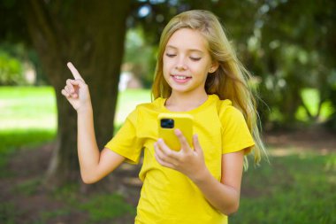 Sarı tişört giymiş, gülümseyen beyaz bir kız dışarıda durup bir elinde telefonu tutan boş bir yeri işaret ediyor.