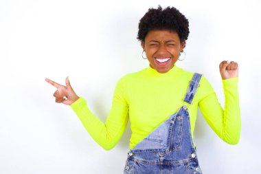 Beyaz duvara karşı kot pantolon giyen neşeli Afro-Amerikan kadın fotokopi reklamı göstererek şansı kutluyor.