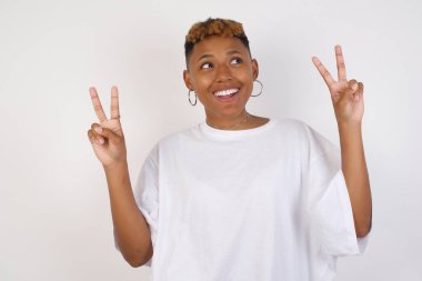 Beyaz tişört giyen neşeli, genç Afrikalı Amerikalı bir kadın iki eliyle barış ya da zafer işareti yapar, rahat kıyafetler giyer, serin hisseder dişli bir gülümsemesi vardır, gri arka planda izole olur. İnsanlar ve vücut dili.